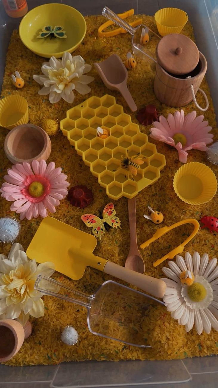 A bee and honeycomb sensory tray with yellow rice, flowers and scoops