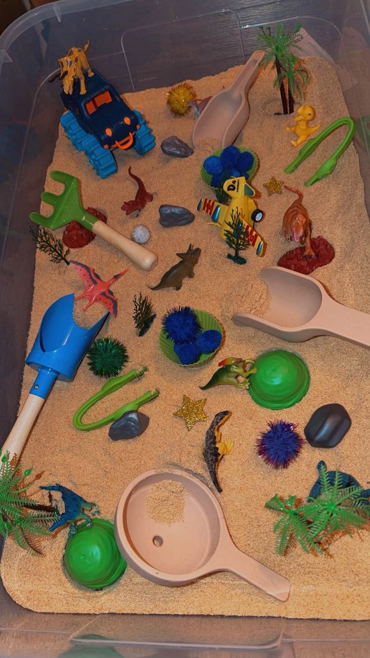 A dinosaur sensory tray with sand, small dinosaurs, palm trees and scoops
